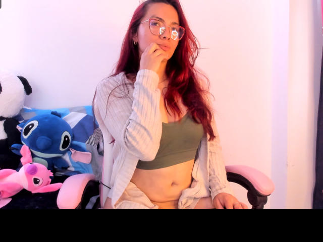 EimmyHoskX with red hair, glasses, green crop top, white cardigan, and loose pants, sitting on a pink chair, hand near mouth, surrounded by blue and pink stuffed animals.