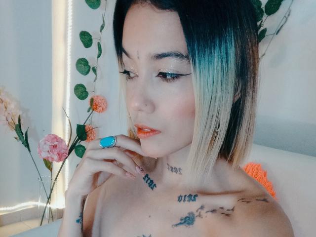Veroo69 with blonde-to-black ombre hair, glittery eyes, orange lipstick, blue ring, tattoos on neck and chest, touching chin, white background with green vines and flowers.