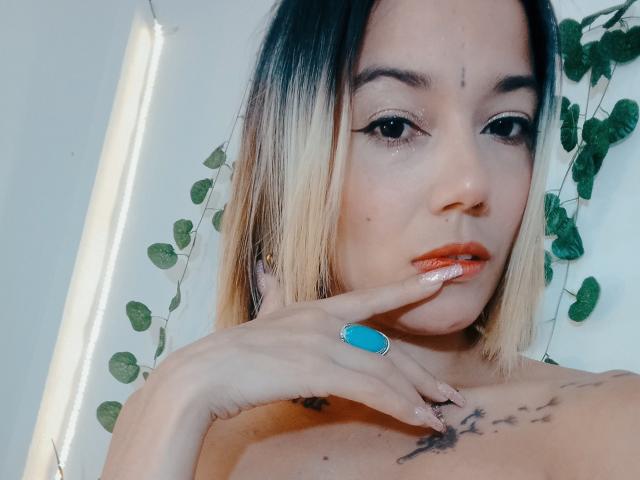 Veroo69 with blonde to black ombre hair, light skin, and tattoos, biting finger with blue ring, orange lipstick, and winged eyeliner, looking at camera.