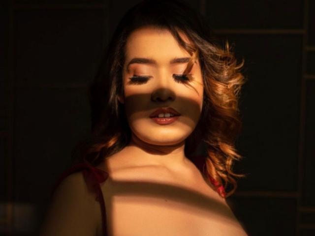 AkiraZanoli with wavy brown hair, closed eyes, and dark lipstick, illuminated by warm sunlight, showing soft shadows on her face and chest. She's wearing a red top.