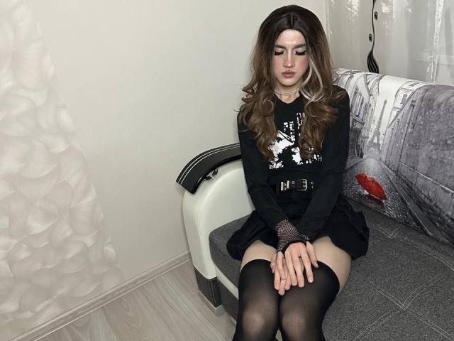 ElyseWhite with long brown hair, wearing a black dress, belt, and fishnet sleeves, sitting with hands on knees, black thigh-highs, in a room with white wall and bed.