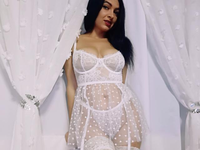 PassionX met lang zwart haar, gekleed in doorschijnende witte kant lingerie, waarbij haar borsten en vulva zichtbaar zijn, staat voor witte gordijnen met bloemen, en glimlacht licht..