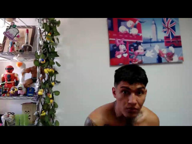 AbrahamStiff with short black hair, shirtless, tattoo on right arm, looking to the side, in a room with a shelf of toys and a colorful painting.