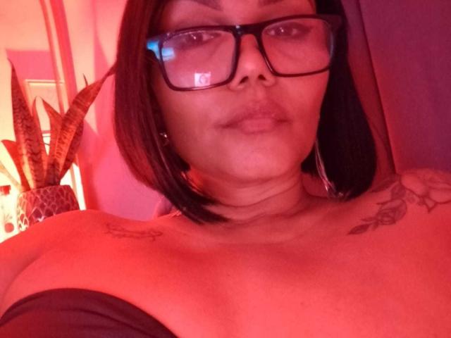 ExoticLyla with red-tinted skin, black shoulder-length hair, and glasses, showing her bare shoulders and tattoos, with her puffy lips slightly parted. Boobs visible, wearing a strapless top.