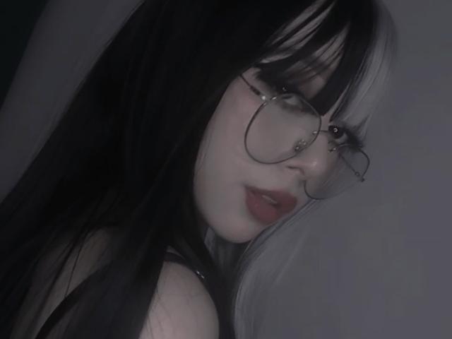 AcidCandy with dark and white hair, wearing glasses, red lipstick, and a black top, looking over her shoulder, showing pale skin and a subtle nose piercing.
