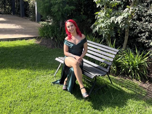 AmyStreetwise69 with red hair, black off-shoulder dress, and green skirt, sitting on a white bench, smiling, showing her tits, on a grassy lawn, wearing silver sandals.