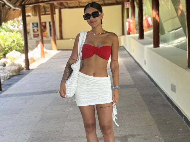 ThaliaAres with dark skin, tattoos, wearing black sunglasses, red strapless top, and white knitted mini skirt, holding a white bag, standing outdoors.