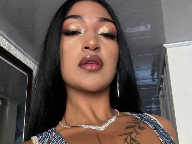 SovyAne69 has glossy golden eyeshadow, long black hair, shiny pink lips, nose ring, silver necklace, and a tattoo on her chest with blue, striped top.