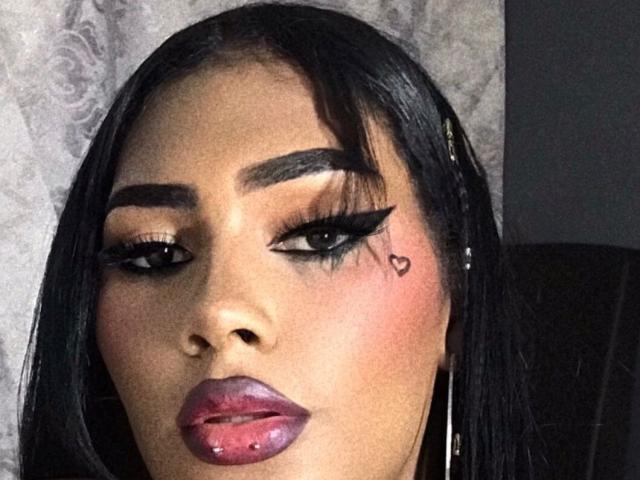 CristinaMontiel has dark, straight hair, thick eyebrows, and dramatic eyeliner with a heart tattoo near her right eye. Her plump, glossy purple lips are slightly parted.