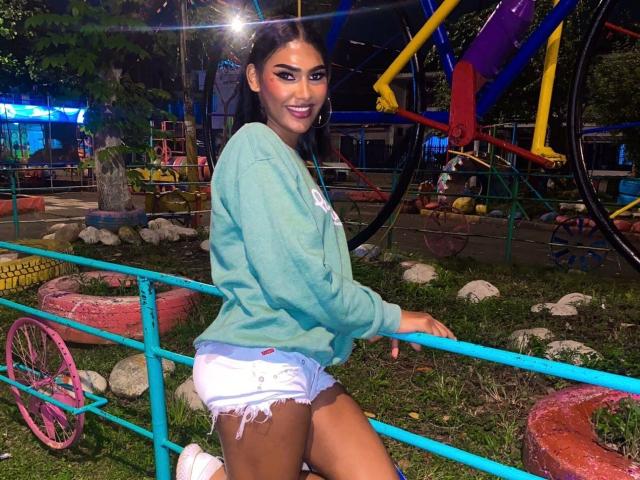 CristinaMontiel with dark hair, green sweater, and white frayed shorts, kneeling by a blue metal fence, smiling at the camera, in a colorful playground at night.