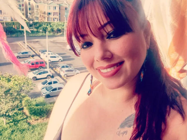 Ccristall has red hair, bangs, and a tattoo on her chest. She's smiling with pink lipstick, wearing colorful earrings, and a strap dress. Her tits are small to medium.