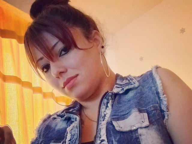Ccristall with red hair in a bun, wearing a frayed denim sleeveless vest, hoop earrings, and a slight smile, showing her cute face and small tits.