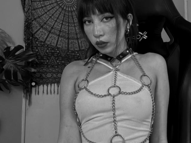 IzzysMisss with dark bangs, wearing a chained harness over her tits, a studded collar, and a ribbed tank top, looking serious in a black-and-white photo.