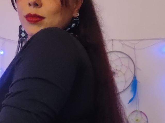 Ccristall's face with red lipstick, black long-sleeve shirt, long brown ponytail, black earrings, and a slight smile, showing a bit of her ass. Soft purple and blue lights.