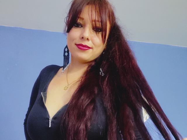 Ccristall has long, dark red hair, wearing a black zip-up top, with purple lipstick and a gold necklace. Her tits are small, and she's smiling slightly.