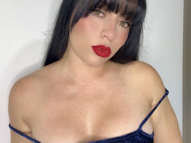 PammelaPrice has dark black hair, red lipstick, green eyes, and a blue velvet top showing her small boobs and fair skin. Her straight hair has bangs.