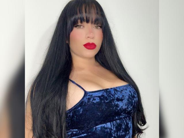 PammelaPrice with long black hair, blue velvet dress, red lipstick, and bangs, showing her tits and a serious expression.