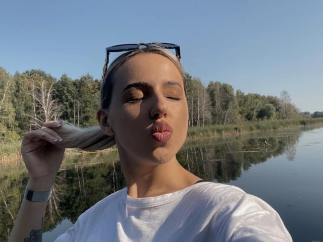 SweetBebea with eyes closed, puckering lips, holding blonde hair, wearing white tee, sunglasses on head, reflection in water, clear blue sky, forest in background.