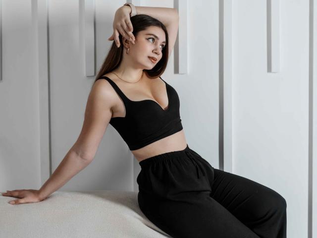 MilanaKitty, avec une poitrine de taille moyenne, un soutien-gorge de sport noir et un pantalon noir, assise sur un banc blanc, bras levé, cheveux bruns, peau claire, sourire discret, regardant sur le côté..