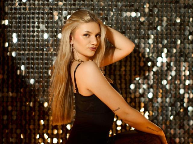 SelenaeMoore met lang blond haar, een zwart tanktopje en een subtiele tatoeage op haar rechterarm, poseert met haar hand in haar haar, waarbij haar achterkant zichtbaar is, en ze een zelfverzekerde glimlach heeft..