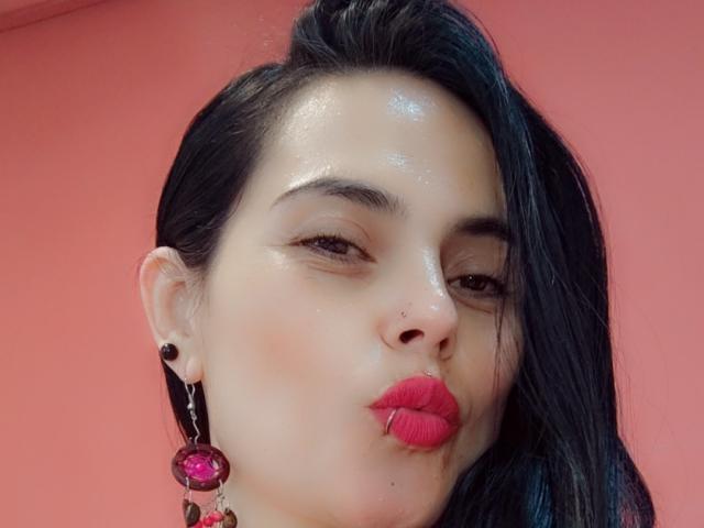 CharlotteCrofort tem a pele brilhante, cabelo preto, batom vermelho e um piercing no nariz. Ela está fazendo beicinho com os lábios rosados, usando alargadores de orelha pretos e um brinco rosa com uma pedra..