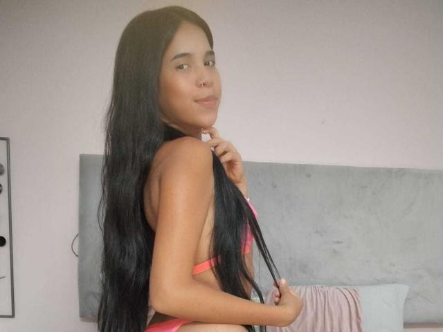 MarySmiith, con cabello largo y negro, vestida con lencería roja, sonriendo a la cámara, sosteniendo su cabello, mostrando su espalda y trasero, en un dormitorio con un cabecero gris..
