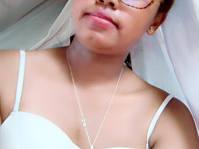 Goddess69s, con la pelle marrone chiaro, che indossa occhiali e un reggiseno bianco, ha piccoli seni e una collana d'argento, con un leggero sorriso, con un tessuto bianco sullo sfondo..