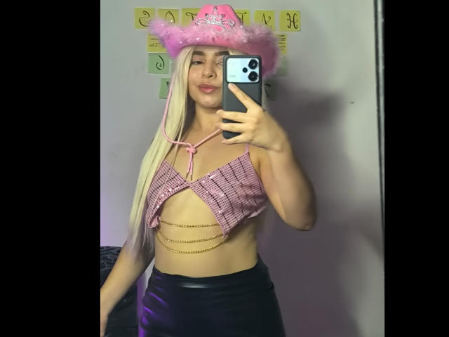 LissyCarter with long blonde hair, pink cowboy hat, pink plaid top showing her tits with chain details, black leather skirt, taking a mirror selfie with an iPhone.