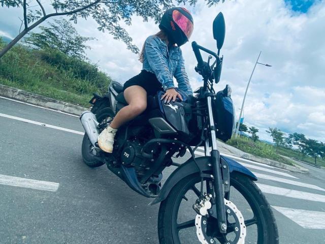 AilenTovar on a black motorcycle, wearing a blue denim jacket, black shorts, yellow sneakers, and a pink helmet, riding on a road with a crosswalk.