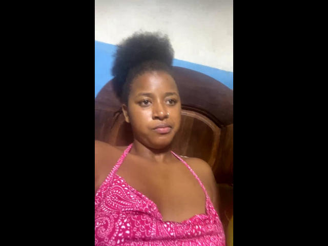 Laulah with dark skin, small afro, wearing a pink halter top with a pattern, has a serious expression, medium-sized tits, and is looking slightly to the left.