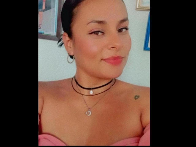 LunaSoler has light brown skin, black hair in a bun, small hoop earrings, black choker, pink off-shoulder top, red lipstick, subtle makeup, small heart tattoo on right shoulder.