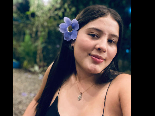 KathyStone met een schattige paarse bloem in haar bruine haar, gekleed in een zwarte top, met een zachte glimlach, waarbij haar kleine, stevige borsten zichtbaar zijn, en een delicate ketting met een klein hangertje..