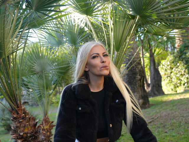BellaTwine with long blonde hair, black jacket, and black top, standing under green palm trees, looking to the side with a slight pout, sunlight filtering through.