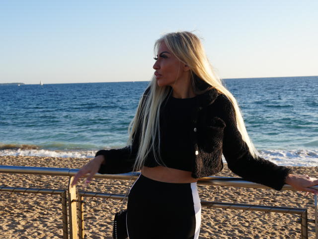 BellaTwine with long blonde hair, black fluffy jacket, black and white pants, standing with arms on beach railing, looking sideways, ocean and clear sky in background.
