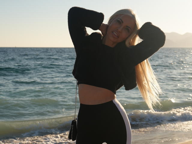 BellaTwine with long blonde hair, black crop top, and black-and-white pants, hands in hair, standing by the ocean, sun setting, small black purse with chain.