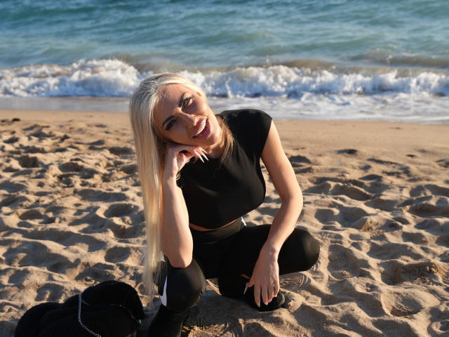 BellaTwine with blonde hair, black top, and black pants, squatting on sandy beach, hand on cheek, smiling, ocean waves in background, black bag to her left.
