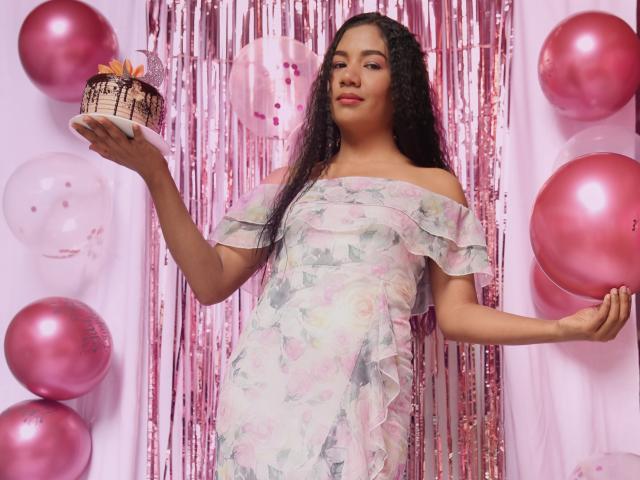 AnaPorto with long curly black hair, pink floral off-shoulder dress, holding chocolate cake with pink frosting, surrounded by pink balloons, pink sequin curtain.