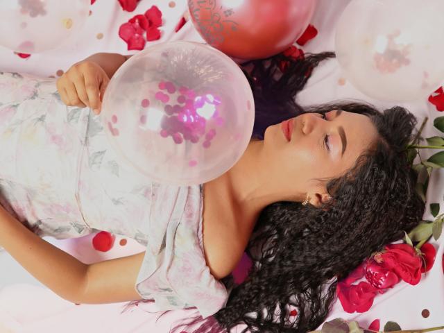 AnaPorto, con i capelli neri ricci, indossa un abito fiorito rosa e sta soffiando un palloncino trasparente con pois viola, circondata da rose rosse e palloncini bianchi, sdraiata su un tessuto rosa..