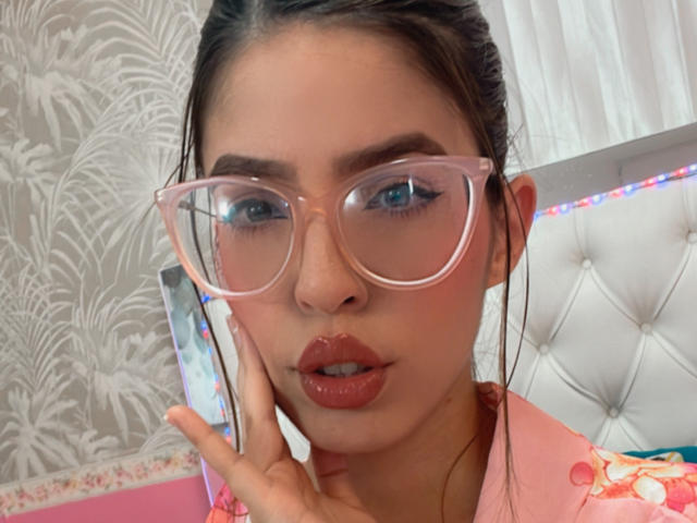 NatyBakers with pink glasses, red lipstick, and pink shirt, holding phone to ear, looking at camera, brown hair in bun, blue eyes, white tufted headboard in background.