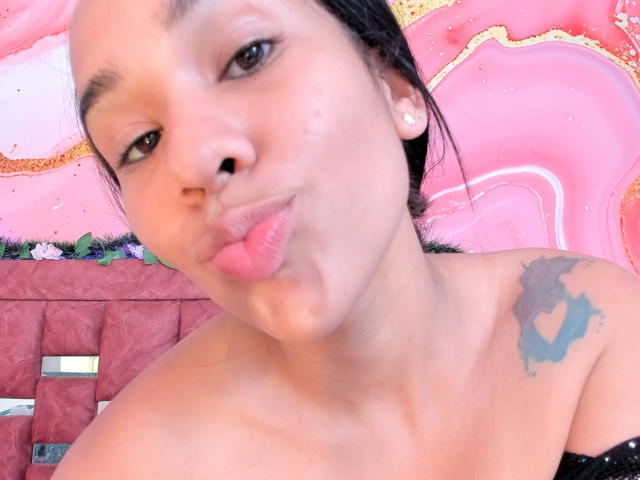 SolStephanny with pouty pink lips, dark brown eyes, and a tattoo on her right shoulder. She's making a kissing face, wearing small white earrings, and has black hair.