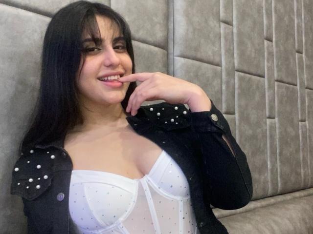 ChloePrie with black jacket, white see-through bustier, showing tits, biting finger, smiling, long black hair, light skin, brown eyes, standing against gray upholstered wall.
