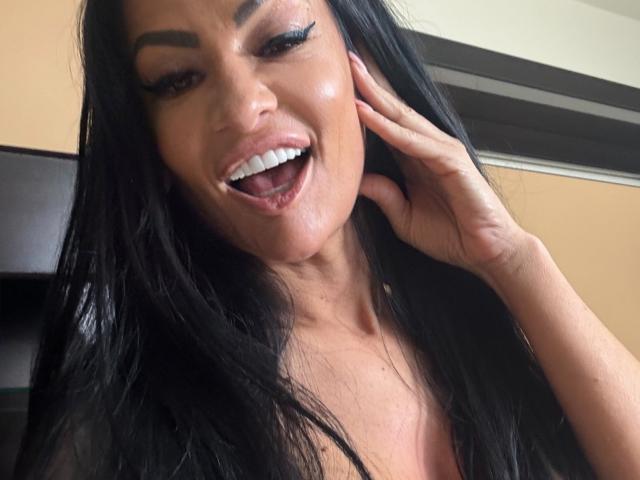 AndraD with long black hair, smiling with her tongue slightly out, showing white teeth, touching her cheek with her right hand, wearing heavy eyeliner and glossy pink lipstick.