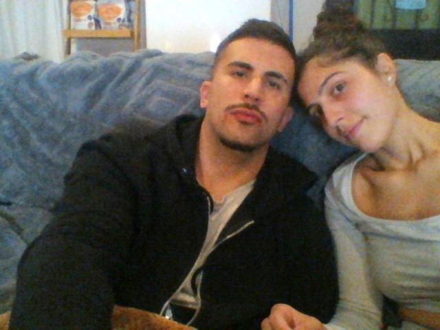 SexyCoupleX69 montre un homme avec une moustache, des cheveux bruns courts et un sweat à capuche noir, à côté d'une fille avec des cheveux bruns bouclés, de petites seins et un haut gris. Ils sont assis sur un canapé..