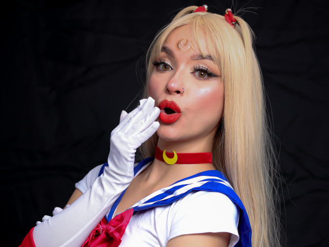 MaryMilan with blonde hair, red lips, white gloves, and blue sailor outfit, biting her finger, red choker, crescent moon, and red hairpins, showing her shiny, cute face.