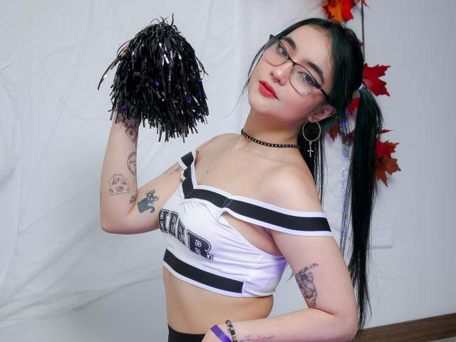 LyRose with green pigtails, glasses, nose ring, choker, and tattoos, holding black pom-poms, wearing a white and black sports bra, and black pants, showing her tits.