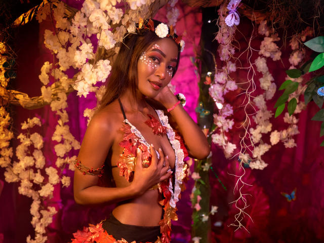 Layla Baker, mit brauner Haut und langen, braunen Haaren, trägt ein Blumenkranz und Körperdekoration, bedeckt ihre Brüste mit den Händen und ist von weißen und rosa Blumen umgeben, lächelnd..