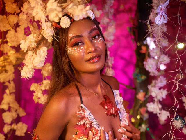 Layla Baker, con piel morena, un tocado floral, lunares blancos en la cara, sombra de ojos rosa, labios brillantes, pechos cubiertos de encaje, un collar de flores rojas, rodeada de flores blancas y luz rosa..