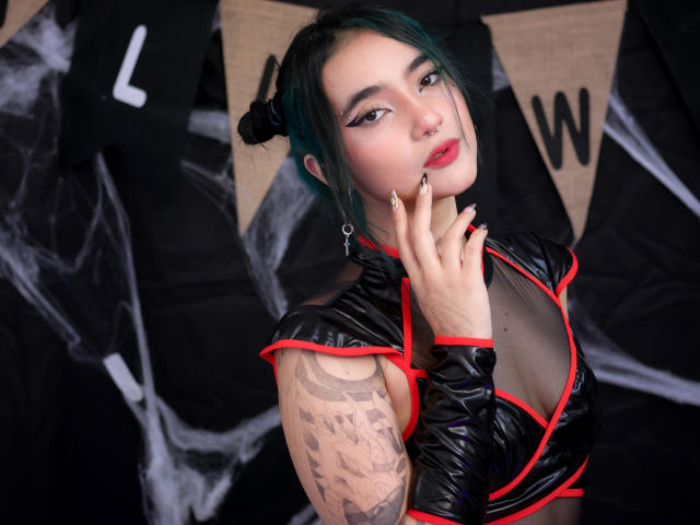 LyRose with green hair, black latex top, red trim, tattoos on right arm, nose ring, red lipstick, hand on chin, looking at camera, black and white background.