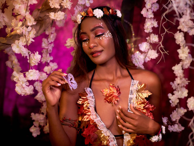 Layla Baker, mit dunkler Haut, trägt ein grünes, offenes Oberteil und hält einen weißen Schmetterling. Ihr Gesicht ist mit weißen Punkten verziert, sie trägt einen Blumenkranz. Im Hintergrund sind rosa und violette Blumen zu sehen, die herabhängen..
