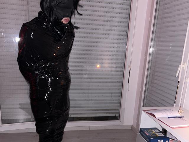 A DominaCaty está envolta em plástico preto brilhante, com os olhos vendados, e está em pé em um quarto. Ela está amarrada com os braços atrás das costas, mostrando apenas seus lábios e queixo..